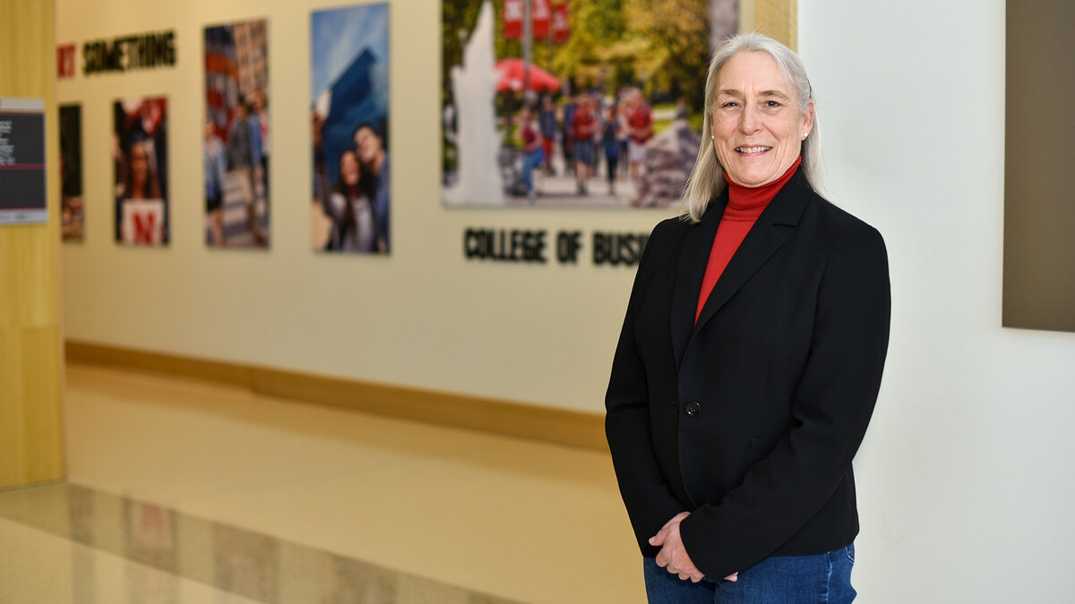 Kay Maresh stands in the College of Business in dark jeans, a red turtleneck and a black blazer. Her hair is long and gray and her hands are put together in her front.